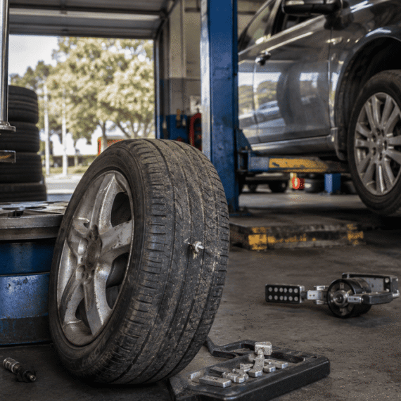 Worn car tyre beside workshop equipment in an Australian tyre service bay, with a vehicle raised on a lift.